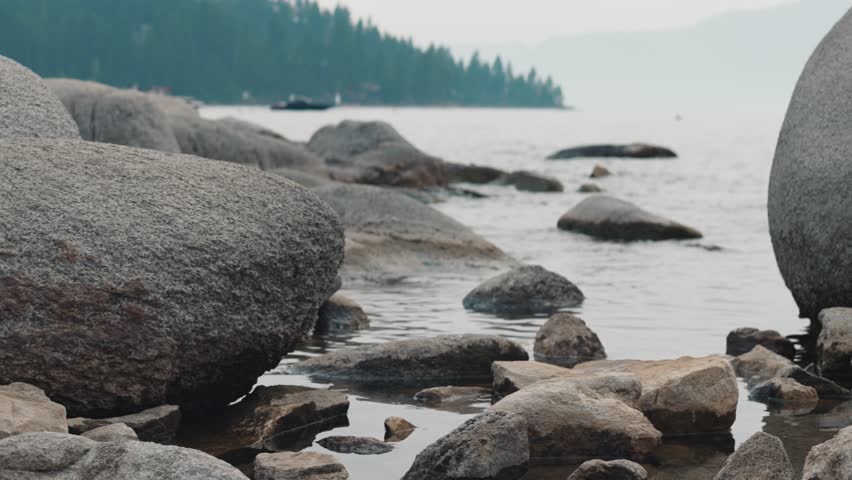 Large boulders and rocks on the shore of calm lake, forested coastline in background with light haze over horizon
