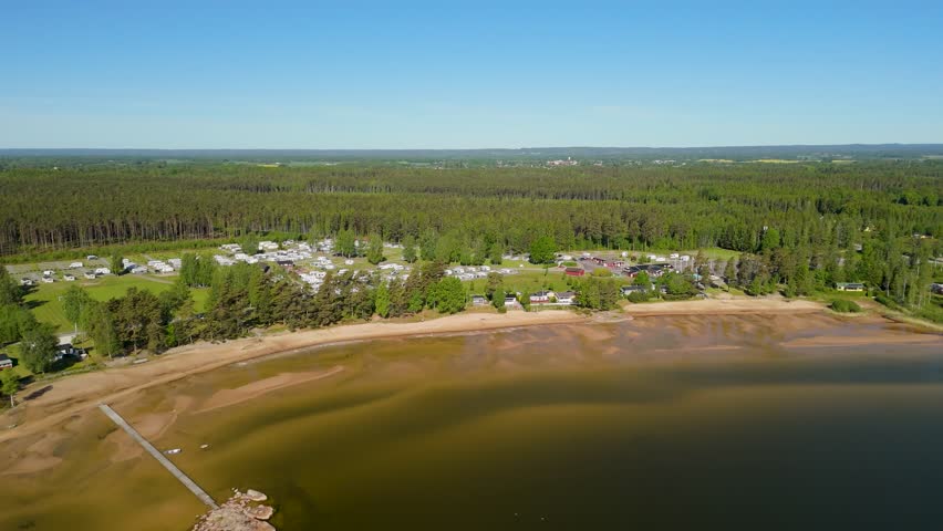 Dramatic aerial pull back shot starting from the wooden pier revealing the long sandy shoreline of Vita Sandar Beach and the nearby camping resort on Lake Vänern in Mellerud Sweden during summer