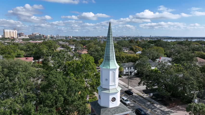 Aerial orbit shot of historic church steeple rising above Hyde Park neighborhood in Tampa, Florida, with green tree canopies. Residential streets and downtown skyline under bright blue sky in distance