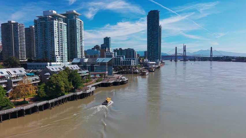 Drone footage of a water taxi traveling along the Fraser River past the downtown skyline of New Westminster, British Columbia, with high-rise buildings and the Pattullo Bridge in the background.