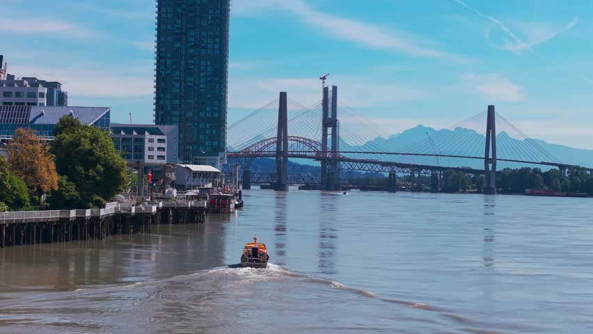 Drone footage of a water taxi traveling along the Fraser River past the downtown skyline of New Westminster, British Columbia, with high-rise buildings and the Pattullo Bridge in the background.