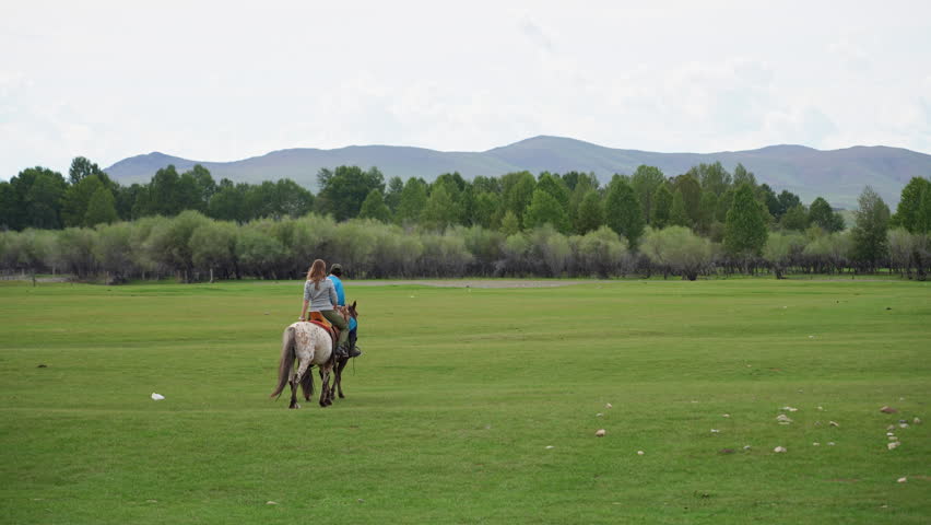 Mongolian Horse Herder Guiding Tourist on Horseback Across Green Steppe Meadow