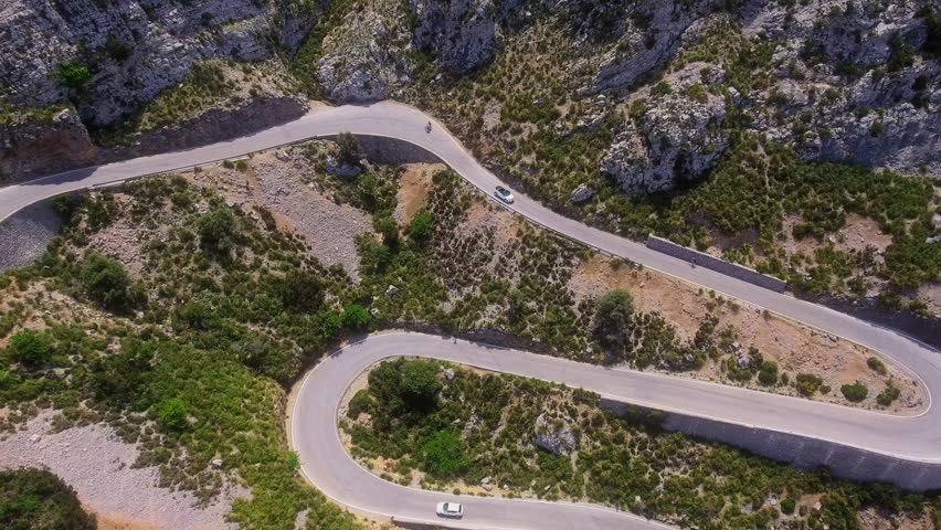 Aerial view shifts to top down as a convertible car turns along a sharp mountain road curve in Mallorca