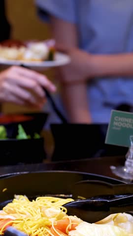 Diner uses tongs to serve fresh noodles and vegetables at bright self-service buffet counter