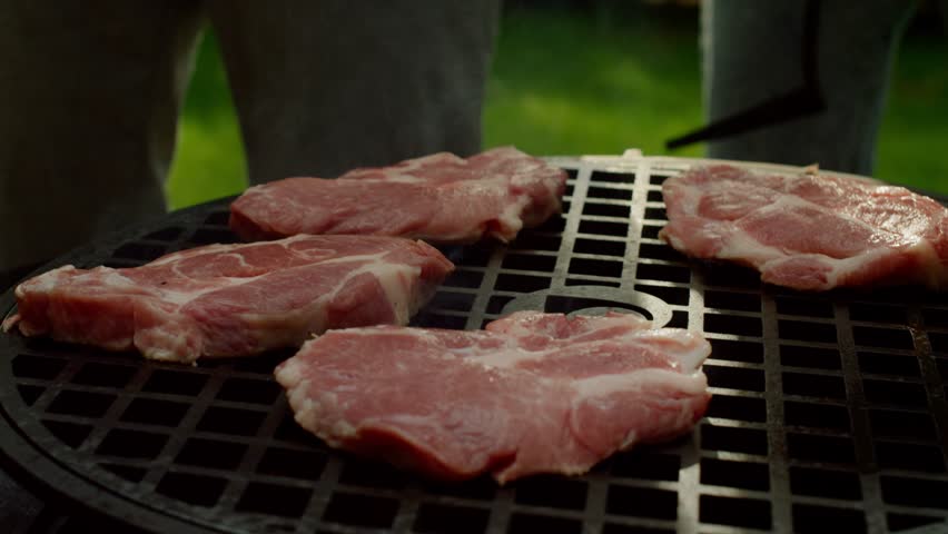 Person grilling pork steaks on outdoor barbecue turning meat with tongs