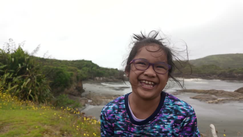 Slow motion footage shows an Asian young girl laughing freely by the beach, with overcast skies and strong breeze, capturing pure joy, childhood happiness, and coastal atmosphere.
