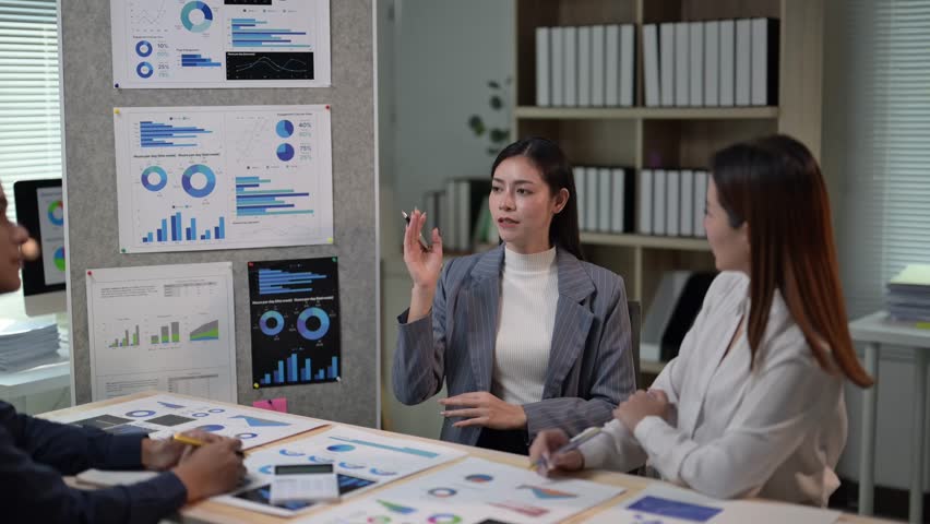 Three women are sitting at a table with a lot of papers and graphs. One of the women is pointing at a graph