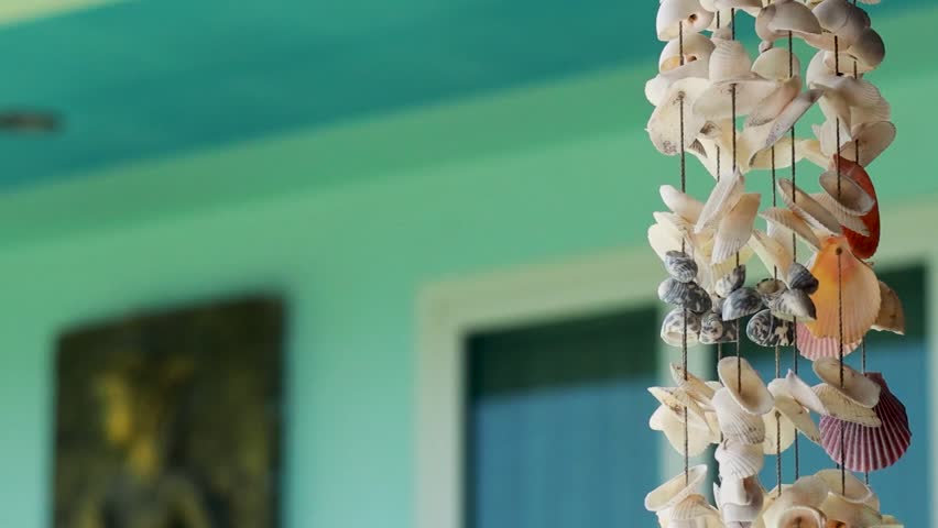 A detailed close-up of a handcrafted shell wind chime swaying against a vibrant teal building.