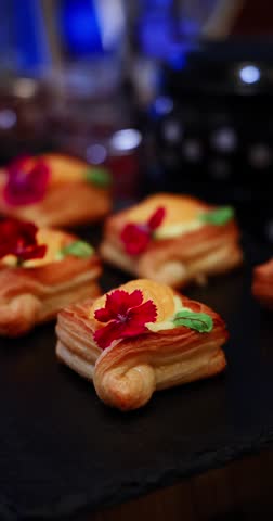 Flaky puff pastries with creamy filling and red edible flower garnish on a dark serving tray. Elegant catering food for events and celebrations.