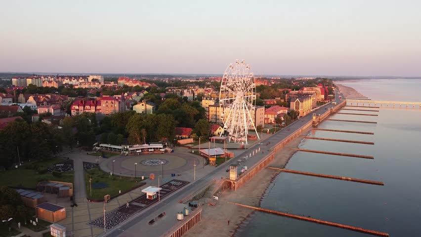 Sunrise Promenade And Ferris Wheel Pastel Dawn With Calm Sea, Empty Boardwalk, Decorative Plaza, Soft Light Bathing Coastal Town And Piers Stretching Into Bay Captured From Above.