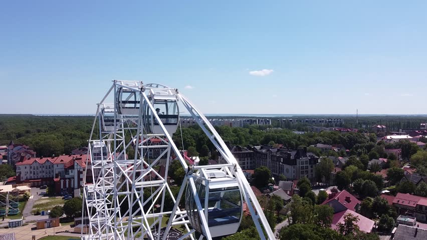 Aerial Daylight Ferris Wheel Over Coastline With Blue Sky, White Wheel Structure Framing Ocean, Sunlit Rooftops, Green Treeline, Beach Promenade And Seaside Resort Atmosphere Captured By Drone.