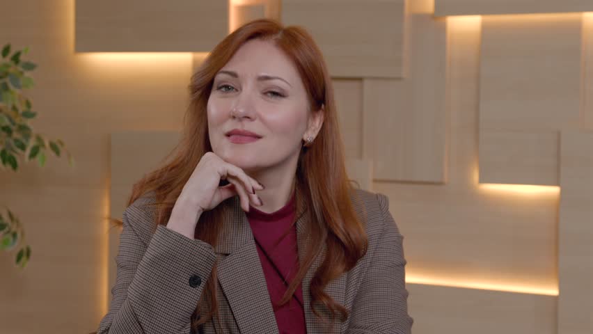 Portrait of charming redhead middle aged woman in stylish business casual looking with friendly smile, expressing confidence and natural beauty while posing at office.