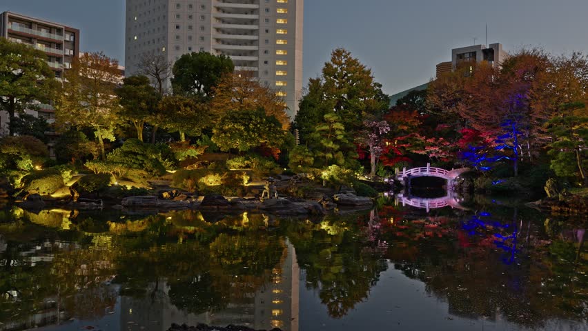 Wide shot of a traditional Japanese garden at night with city buildings and pond reflections.