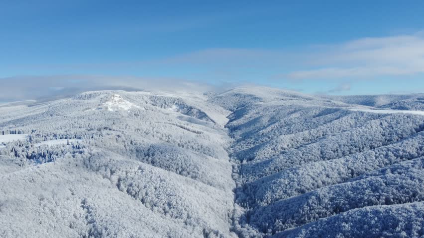 An aerial view of a vast, snow-covered mountain range. A series of rolling, forested mountains covered in a thick blanket of snow