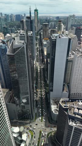 Breathtaking vertical slow-motion aerial perspective smoothly rising upward over the prestigious Makati business district in Manila, Philippines. This cinematic footage begins with ground-level boulevards and gradually ascends to showcase an impressive skyline of contemporary glass-and-steel towers reaching toward overcast heavens. The ascending camera movement reveals multiple layers of commercial real estate, from foreground structures to distant mountain ranges visible on the horizon. Prominent architectural features include sleek corporate headquarters with reflective facades, construction cranes indicating ongoing development, and tree-lined avenues cutting through dense clusters of high-rise buildings. Visible transportation networks show heavy vehicular flow along main thoroughfares and intricate intersection patterns. The atmospheric cloudy conditions create dramatic lighting across concrete surfaces while emphasizing the vertical scale of this Southeast Asian financial hub. Frame composition captures diverse building designs ranging from modern minimalist blocks to ornate residential condominiums. This professional stock content documents the economic heartbeat of the Philippine capital, demonstrating rapid urbanization and metropolitan growth in tropical Asia.