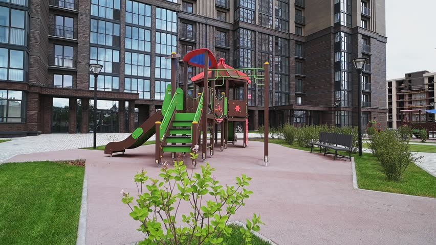A modern children’s playground with climbing frames, a red-roofed playhouse, green handrails, situated near residential buildings, with benches nearby.
