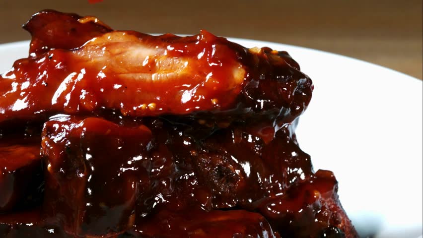 Thick red barbecue sauce being poured over a stack of juicy grilled pork ribs on a white plate in extreme close up
