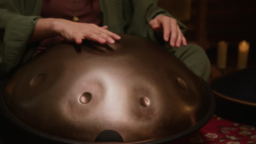 Medium midsection shot of unrecognizable female performer playing hang drum or handpan, while sitting on carpet during sound healing session, esoteric concert or meditation party