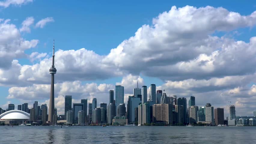 This time-lapse video offers a breathtaking panoramic view of the Toronto skyline set against a vibrant, deep blue sky. The iconic CN Tower stands as a central anchor on the left, while a dense array of modern skyscrapers stretches across the horizon, their glass surfaces reflecting the shifting light. Large, fluffy white clouds drift rapidly across the frame, creating a dynamic sense of movement and changing shadows over the city below. In the foreground, the calm waters of Lake Ontario provide a serene base for the urban energy, with a ferry seen gliding across the harbor, illustrating the seamless blend of metropolitan life and natural beauty.