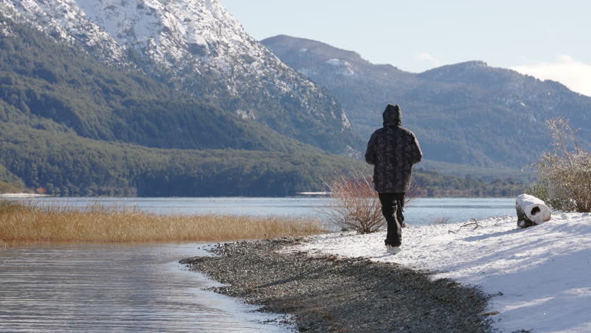 A lone trekker in winter gear navigates a snow-lined lake shore, framed by towering snow-capped mountains and dense alpine forests in Andean or Patagonian landscape.