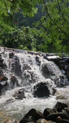 A short clip showing a man-made cascade or small waterfall flowing over a low ledge. The water tumbles rapidly into a pool below, with surrounding trees and greenery creating a lush, park-like setting. The motion is dynamic, capturing the splash and white water as it falls