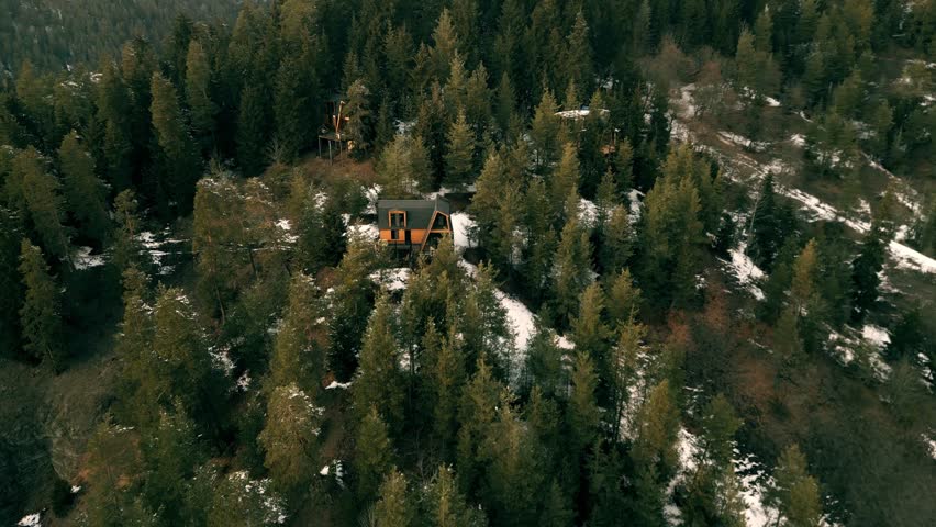 Aerial drone footage showing a small wooden A-frame cabin nestled among dense pine trees on a mountain slope with patches of snow. The drone hovers and slowly rotates, revealing the forest landscape and surrounding cabins. Captured in soft daylight, this winter forest scene conveys remote mountain living and natural tranquility.