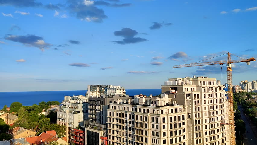 View of building under construction with construction crane, green trees, blue sea and blue sky on sunny summer evening. Roof, windows, walls of new building. City scenery. Urban landscape. Cityscape