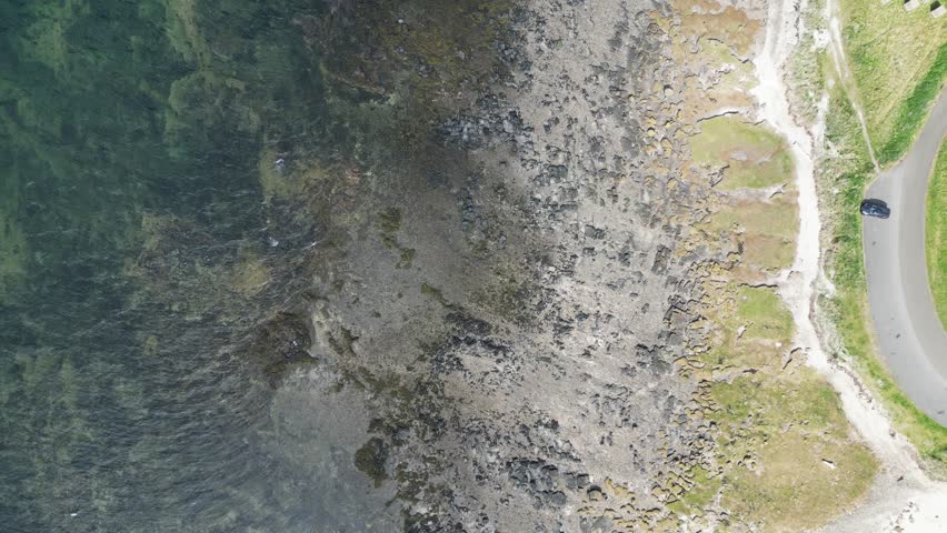 Top-down drone view tracking upward along the shoreline at Longniddry, Scotland, showing rocky coast, sea defences, intertidal water, shallow sea and grassland beside a coastal road.
