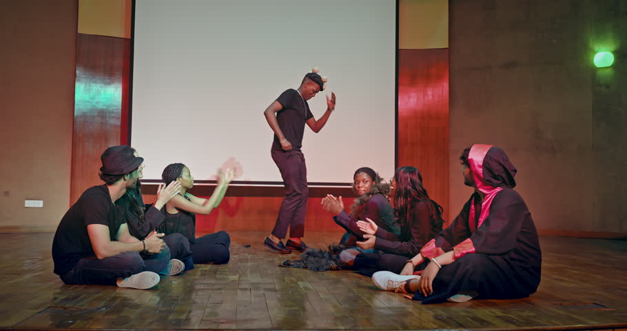 Indian young adult gen z male man wear stage outfit standing do funny dance inside studio hall. Happy smiling teen age boy girl student group team sitting on floor look scene enjoy have fun joy indoor campus place