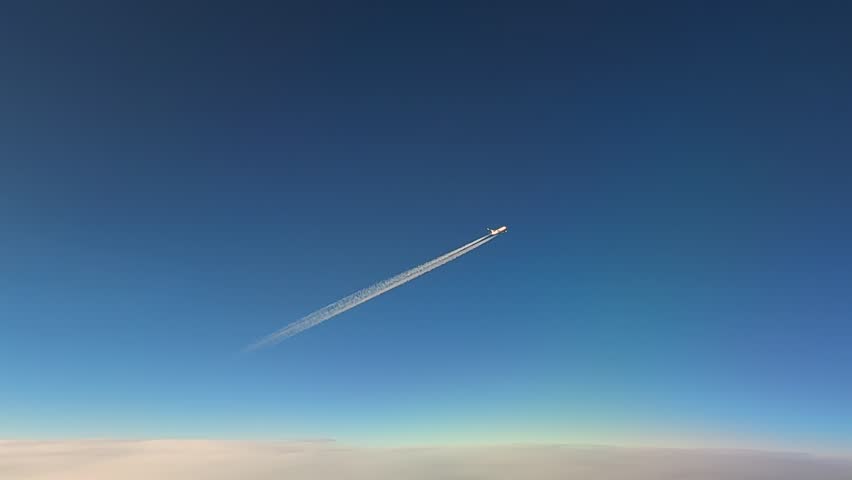 High-angle cockpit view of a commercial jet and its fading contrail fading in a blue sky. Diagonal shot taken from another jet cockpit flying bellow.