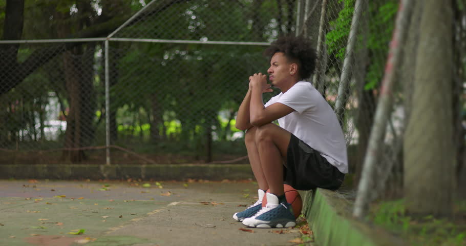 African American young man sitting on outdoor basketball court leaning against fence, deep in thought, reflective moment showing focus, patience, and quiet determination