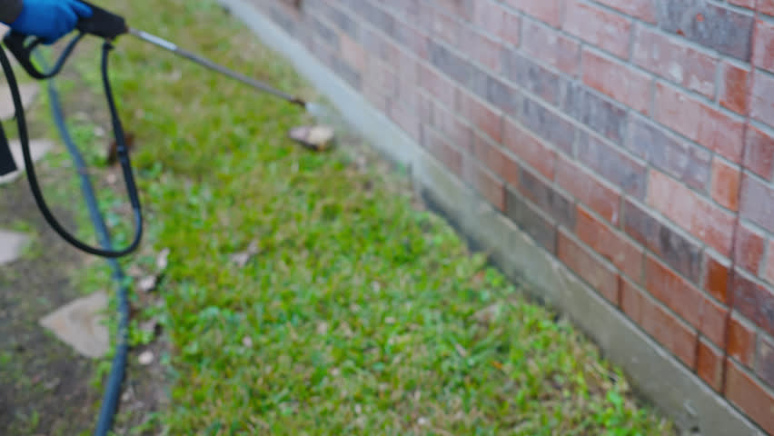 Exterior pest management spraying along house foundation and grass edge, wetting side of wall