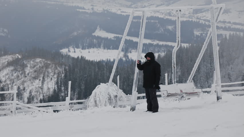Man in black clothes taking photo with smartphone near frozen metal swing on top of snowy mountain with scenic view
