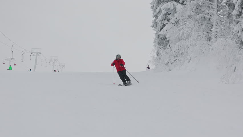 Skier in red jacket and helmet riding down groomed snowy slope at mountain ski resort during cloudy winter day