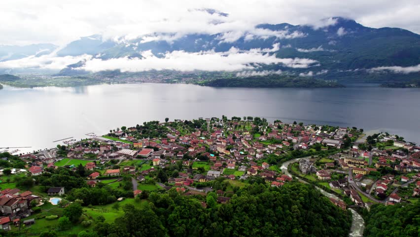 Cinematic aerial drone footage flying backward over a historic city with red rooftops, revealing Lake Como and a large alpine mountain in the background.