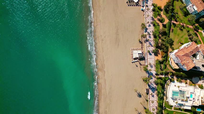 Top‑down flyover of Puerto Banús beach, Marbella, Spain, white sand, turquoise water, five star hotels, pedestrians along the promenade, and La Victoria statue.