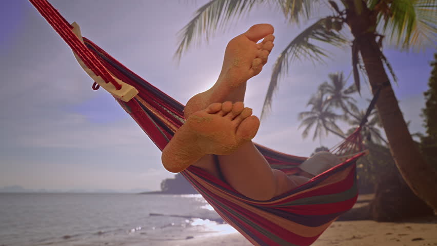 Bare Female Feet Covered With Sand Resting In Hammock Above Tropical Beach With Palm Trees And Soft Evening Light.