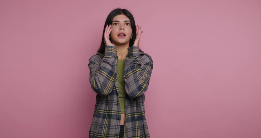 A person with long dark hair shows surprise with hands on their face. They are wearing a plaid shirt over a green top. The background is bright pink, creating a vibrant look.