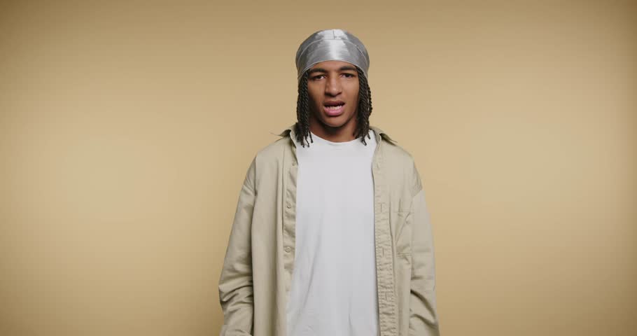 A young man raises his hand to wave while smiling. He is wearing a simple outfit and stands in front of a plain background. The lighting is bright suggesting daytime.