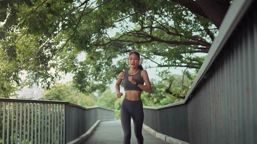 Young fit Asian woman listening music jogging in a park, pausing to check her performance, time, heart rate, pulse, SPO2on a smartwatch before continuing her run
