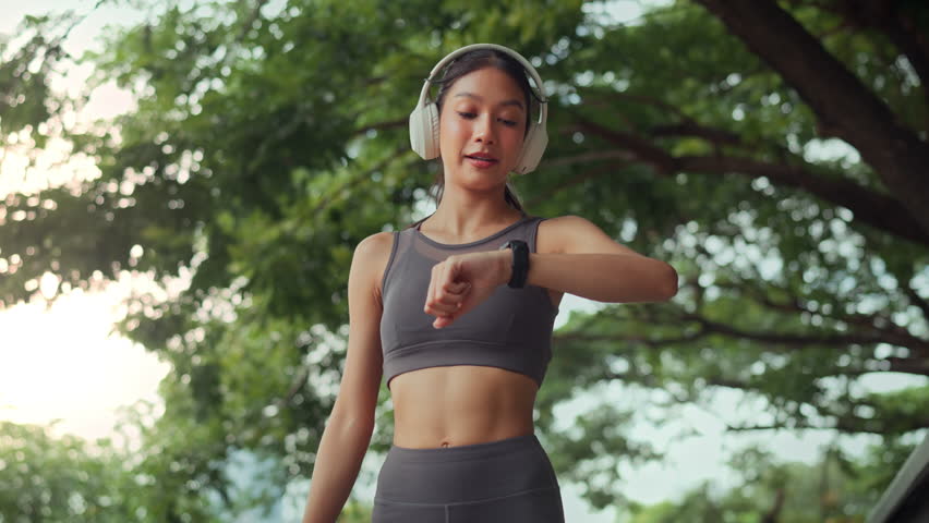 Young fit Asian woman listening music jogging in a park, pausing to check her performance, time, heart rate, pulse, SPO2on a smartwatch before continuing her run