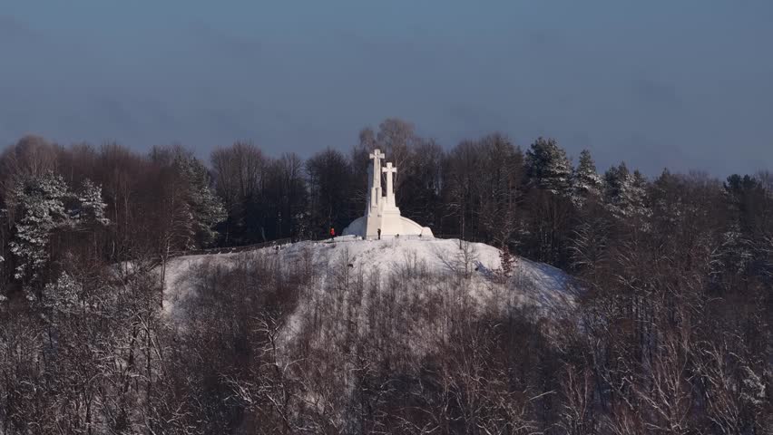 Winter view of the Three Crosses Monument in Vilnius, standing atop a snow-covered hill surrounded by frosty forest under a clear blue sky.