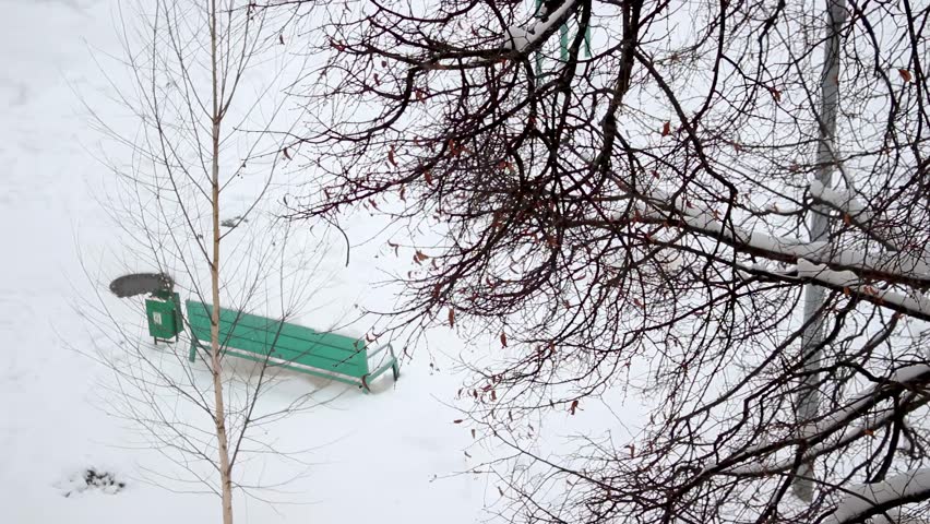 Heavy snowfall and a winter landscape with a green bench covered in snow under bare tree branches