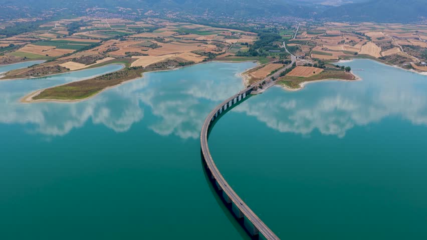 Aerial view of Lake Polyfytos in Greece with a curved bridge, blue waters, and scenic mountains. A peaceful landscape blending nature, architecture, and rural charm