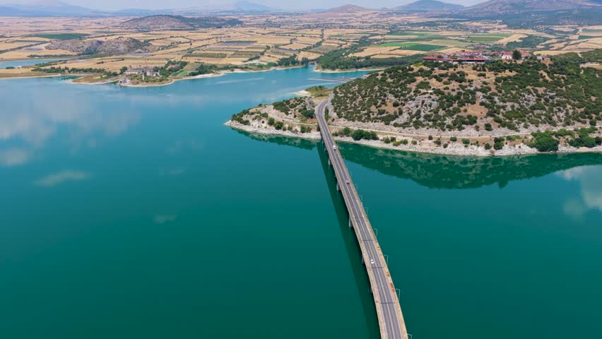Aerial view of Lake Polyfytos in Greece with a curved bridge, blue waters, and scenic mountains. A peaceful landscape blending nature, architecture, and rural charm