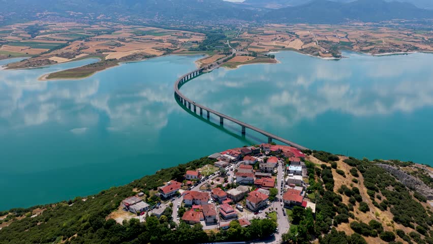 Aerial view of Lake Polyfytos in Greece with a curved bridge, blue waters, and scenic mountains. A peaceful landscape blending nature, architecture, and rural charm