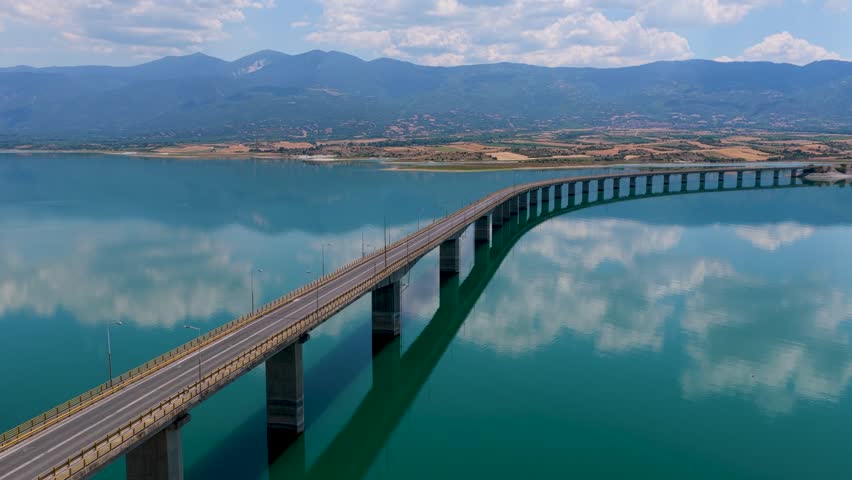 Aerial view of Lake Polyfytos in Greece with a curved bridge, blue waters, and scenic mountains. A peaceful landscape blending nature, architecture, and rural charm