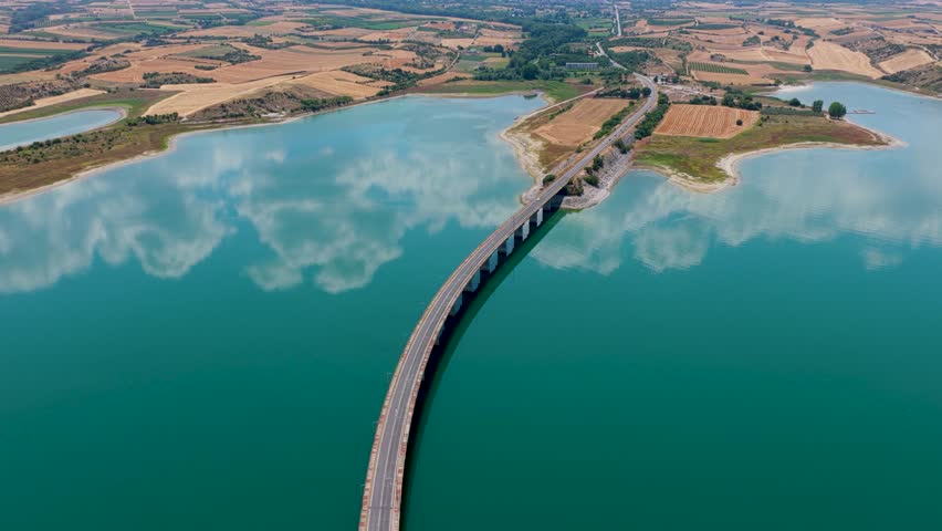 Aerial view of Lake Polyfytos in Greece with a curved bridge, blue waters, and scenic mountains. A peaceful landscape blending nature, architecture, and rural charm
