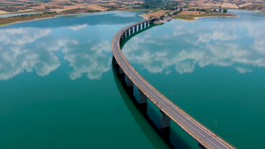 Aerial view of Lake Polyfytos in Greece with a curved bridge, blue waters, and scenic mountains. A peaceful landscape blending nature, architecture, and rural charm