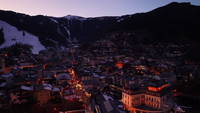 twilight alpine town under snow slopes with glowing streetlights and lit rooftops, ski runs visible as pale ribbons, aerial drone